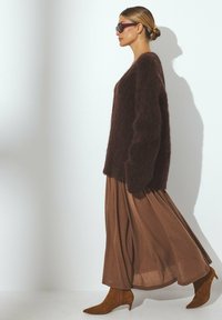Brūns pūkains džemperis, garš brūns svārki un norādīto brūnu zamšādas zābaku. Modelis stāv sāniski, valkājot tumšas saulesbrilles, metot mīkstu ēnu.
