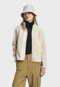 Chaqueta de forro polar en color crema con cierre de presión y cuello alto; llevada sobre un suéter de cuello alto negro. Combinada con pantalones cargo en color beige y un sombrero de cubo de color claro.