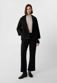 Giacca esterna nera con texture, con rever a punta, indossata su un dolcevita grigio chiaro, abbinata a pantaloni neri e stivaletti.