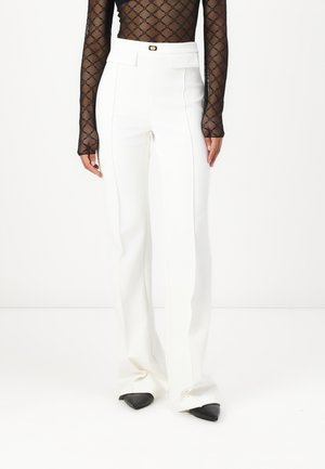 Pantaloni bianchi alla vita alta, sartoriali, con un design a gamba dritta, caratterizzati da una leggera lucentezza e scarpe nere con punta affilata. Sotto, una maglietta nera a fantasia.
