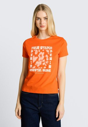 Frau trägt ein orangefarbenes kurzärmeliges T-Shirt mit weißem abstraktem Text und dunkelblaue Jeans, steht vor einem einfarbigen grauen Hintergrund.