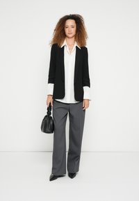 Μαύρο σακάκι, λευκό πουκάμισο, γκρι παντελόνι με φαρδιά μπατζάκια και μαύρες μυτερές γόβες. Κρατάει μια μικρή μαύρη τσάντα με υφή. Ογκώδη σγουρά μαλλιά.