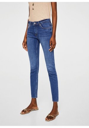 Person iført mid-rise slim fit blå jeans kombineret med beige ærmeløs top og beige stroppesandaler med flad hæl på lys baggrund.