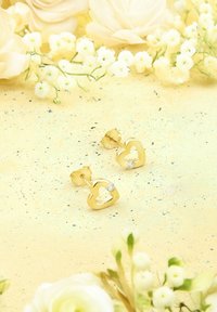 Guld, hjärtformade örhängen med en utstansad design och små klara stenar som dekoration. Mot en mjuk gul bakgrund med blommor.