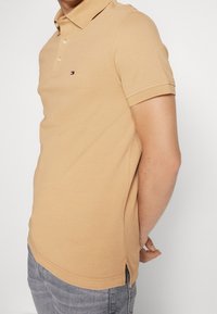 Beżowa koszulka polo z teksturowanej tkaniny, z kołnierzykiem, krótkimi rękawami i małym logo na piersi. Po bokach rozcięcia przy dolnym brzegu.