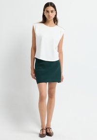 Vrouw die tegen een effen achtergrond staat, gekleed in een mouwloze witte top met pareldetails, een donkergroene minirok en bruine T-strap schoenen.