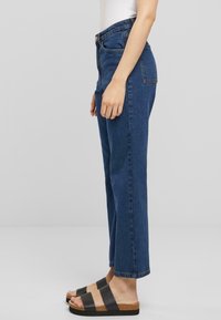 Jeans de tiro alto y pierna recta en denim azul oscuro, con cinco bolsillos y un acabado ligeramente deslavado. Combinados con sandalias negras tipo slide.