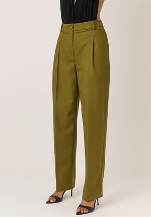 Vrouw draagt olijfgroene, op maat gemaakte broek met hoge taille en plooien, gecombineerd met zwarte open-toe hoge hakken, staand tegen een lichte achtergrond.