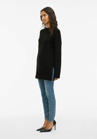 Top de malha preto de manga longa com fendas laterais, emparelhado com jeans azuis justos e sapatos de salto agudo pretos. Textura suave, ajuste relaxado.