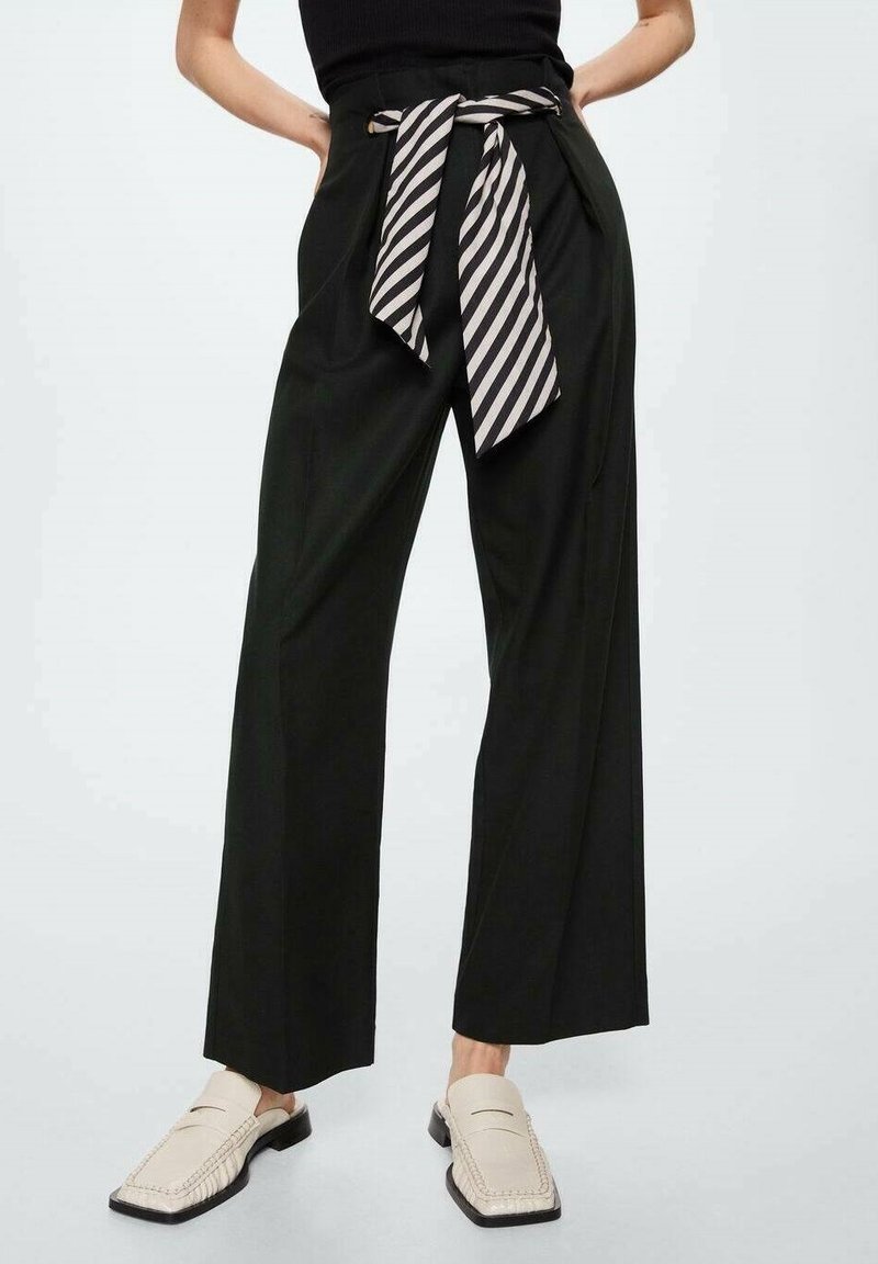 Pantalon noir à jambes larges avec une ceinture rayée noir et blanc nouée à la taille, porté avec des mocassins beige à bout carré sur un mannequin.