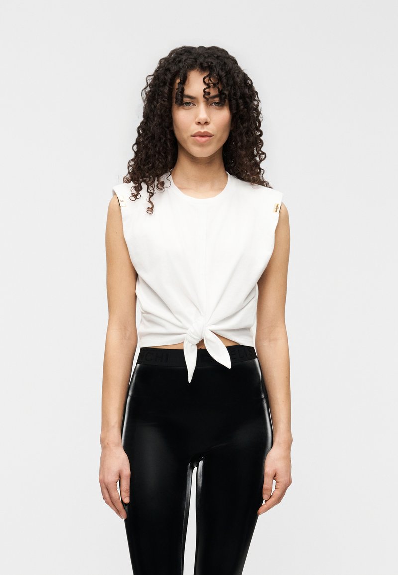 Λευκό crop top με κοντά μανίκια και λεπτομέρεια κόμπου μπροστά. Συνδυάζεται με μαύρο, γυαλιστερό, ψηλόμεσο κολάν. Μινιμαλιστικός σχεδιασμός με χρυσές λεπτομέρειες.