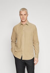 Chemise beige à manches longues en tissu texturé avec un col boutonné, dotée d'une patte de boutonnage à l'avant et d'une coupe décontractée, associée à un pantalon noir.