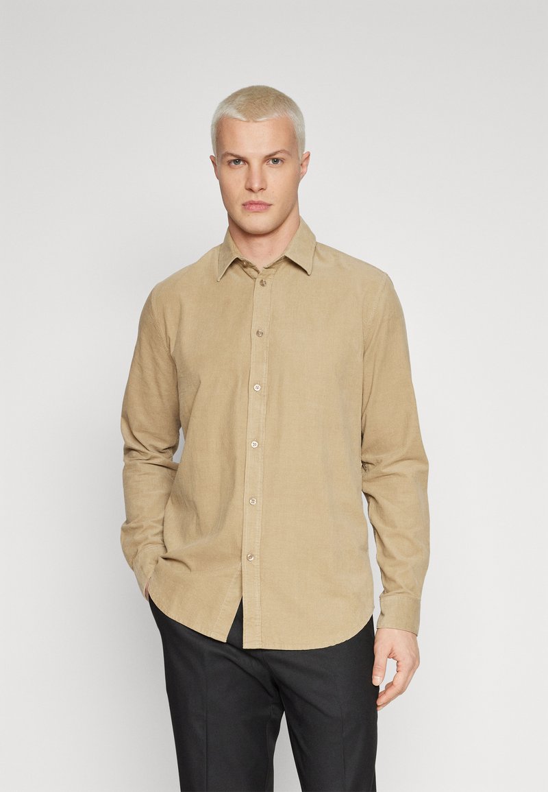 Chemise beige à manches longues en tissu texturé avec un col boutonné, dotée d'une patte de boutonnage à l'avant et d'une coupe décontractée, associée à un pantalon noir.