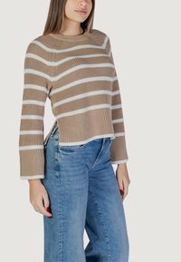 Maglione beige a righe con linee orizzontali bianche, polsini e orlo a coste, con dettagli a fessura laterale. Indossato con jeans a gamba larga blu chiaro.