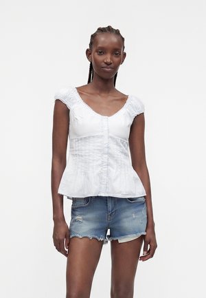 Femme aux cheveux tressés portant un chemisier blanc à manches courtes boutonné et un short en jean usé, debout devant un fond blanc.