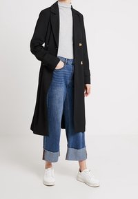 Kvinna klädd i en lång svart kappa med guldknappar, grå polotröja, uppvikta blå jeans och vita sneakers, stående med ena handen i fickan.