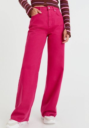 Vrouw draagt felroze, wijde broek met hoge taille, een bruin gestreept top met lange mouwen en witte sneakers, staat met één hand in de zak.