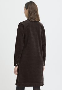 Žena s ramena dužine smeđom kosom, odjevena u dugu tamnosmeđu haljinu od manšetrastog materijala, okrenuta leđima prema jednostavnoj svijetloj pozadini.