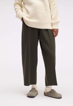 Person wearing cream knit sweater, dark wide-leg pants, beige socks, and taupe Birkenstock clogs standing on a white floor.