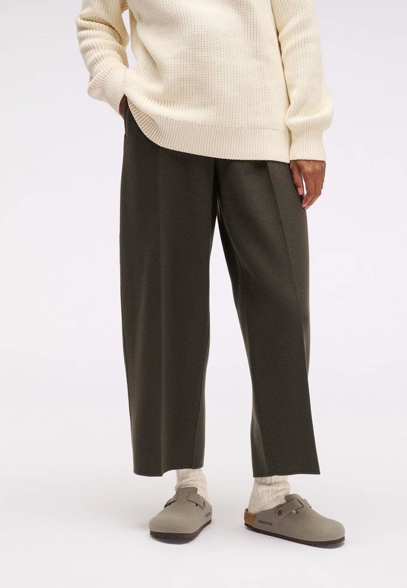 Personne portant un pull en maille crème, un pantalon large foncé, des chaussettes beiges et des sabots Birkenstock taupe, debout sur un sol blanc.