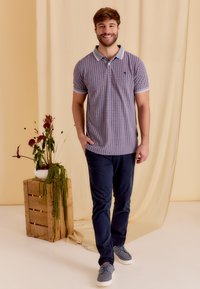 Heren polo met geometrisch patroon in blauw en rood, korte mouwen, knoopsluiting, gecombineerd met marineblauwe broek en grijze sneakers.