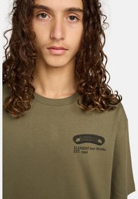 Oliven grøn bomulds t-shirt med rund halsudskæring, korte ærmer, prydet med et sort grafisk tryk med tekst på venstre brystområde.