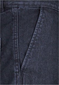 Tejido de mezclilla azul oscuro que muestra una textura suave, con un frente plano y bolsillos en ángulo, y costuras visibles a lo largo de las uniones.