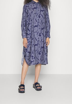 Femme portant une robe violette à manches longues, boutonnée jusqu'au genou, avec des motifs floraux foncés, ainsi que des sandales à plateforme noires à boucles.