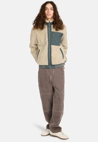 Fleecejakke i creme og mørk teal, med lynlås lukning, kontrastlomme, parret med løse brune bukser og hvide sneakers. Tekstureret yderstof.