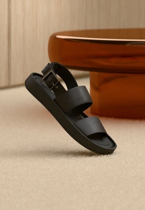 Sort sandal med dobbelte stropper og spændelås på beige tæppe, ved siden af et rundt, ravfarvet bord i et neutralt rum.