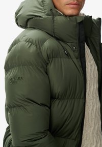 Hombre con chaqueta de plumas verde con capucha y cremallera, superpuesta sobre un suéter de punto trenzado color crema, primer plano del torso superior y la mitad inferior de la cara.