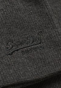 Tejido de punto gris oscuro con una textura acanalada y un sutil logotipo bordado en la esquina inferior izquierda.
