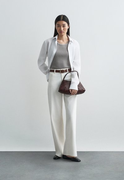 Chemise blanche à manches longues, débardeur rayé gris, pantalon blanc à jambes larges, ceinture marron, sac à main tissé foncé, ballerines noires sur un sol gris.