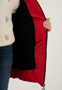 Rød frakke med indvendig foring af sort fleece, med lynlås lukning. Yderskallen er glat, og indersiden har en blød tekstureret overflade.