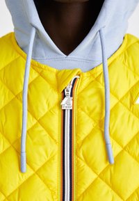 Primo piano di una persona che indossa una giacca trapuntata gialla con una cerniera a righe e un hoodie azzurro chiaro sotto.