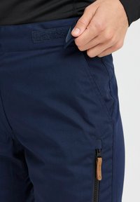 Navy blue hiking pants with a textured fabric, featuring a side zip pocket and a Velcro closure at the waist for secure fit.