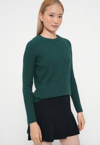 Pull en hiver vert en maille, à manches longues, encolure ronde, finition texturée, associé à une jupe plissée noire. Design simple et ajusté.