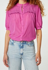 Korte mouwen, knoopsluiting blouse in felroze met een geborduurde, getextureerde schouderpas. Draagt bij hoge taille lichtblauwe jeans met een vervaagd accent.