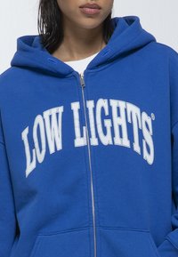 Felpa blu con zip in materiale morbido. Presenta un grande testo bianco "LOW LIGHTS" curvato sopra il petto e tasche frontali. Cappuccio regolabile.