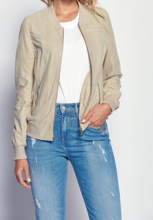 Femme portant une veste beige perforée sur une chemise blanche et un jean bleu déchiré, main dans la poche, sur fond uni.