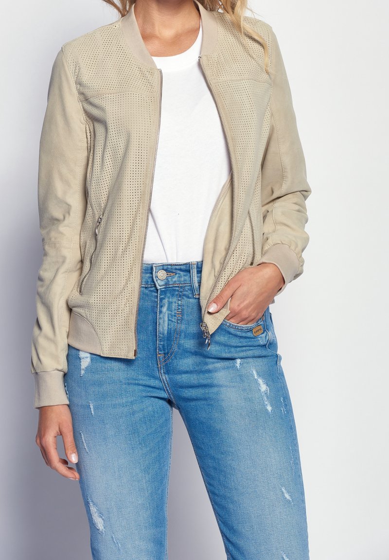 Femme portant une veste beige perforée sur une chemise blanche et un jean bleu déchiré, main dans la poche, sur fond uni.