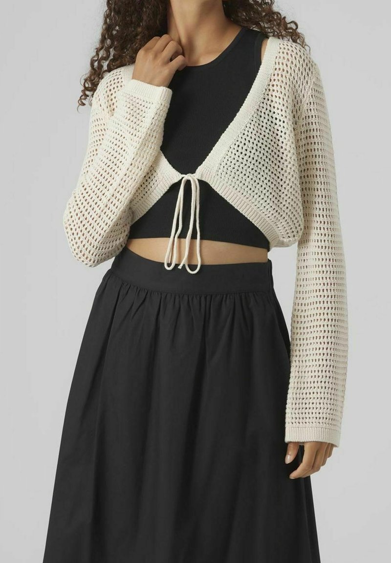 Žena v černém crop topu a černé sukni s vysokým pasem, s krémovým otevřeným pleteným kardiganem svázaným vpředu, pózuje na jednobarevném pozadí.