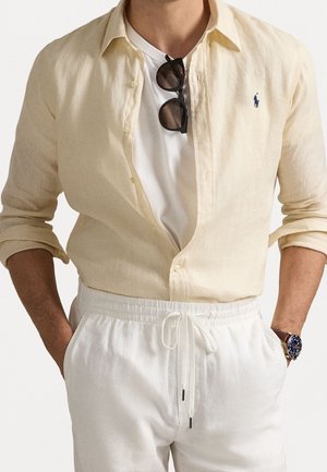 Homme portant une chemise beige clair à boutons par-dessus un t-shirt blanc, un pantalon blanc à cordon, des lunettes de soleil foncées suspendues à la chemise et une montre au poignet gauche.