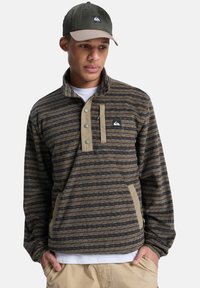 Καφέ φλις πουλόβερ με σχέδιο, υψηλό γιακά, κουμπωτή λωρίδα και αντίθετη τσέπη σε μπεζ χρώμα. Το καπέλο έχει σκούρο πράσινο εμπρός και ανοιχτό καφέ γείσο.