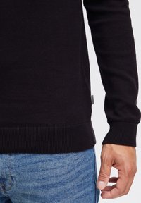 Manche de pull en tricot côtelé noir montrée sur du denim bleu. Présente un ourlet droit et une étiquette latérale avec un motif rayé.