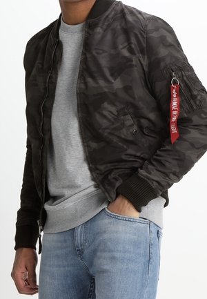 Mężczyzna ubrany w czarną bomberkę moro z czerwoną metką "Remove Before Flight" na rękawie, szarą bluzę i jasno-niebieskie dżinsy.