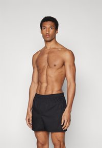 Schwarze Badehose mit elastischem Bund, glatter Textur, knielang, getragen von einem muskulös gebauten Mann, der vor einem schlichten Hintergrund steht.