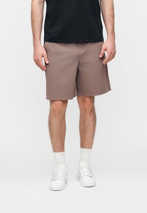 Mees, kes kannab beeži toonis põlveni ulatuvaid lühikesi pükse, valgeid sokke, valgeid tenniseid ja musta särki, seisab ühevärvilise tausta ees.