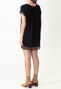 Vestido túnica negro con mangas cortas, que presenta un diseño fruncido en la parte posterior y un dobladillo con encaje negro. La modelo lleva botas de tobillo color marrón.