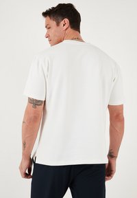 Camiseta blanca de manga corta con escote redondo y aberturas laterales. Textura suave; presenta tatuajes visibles en el brazo.